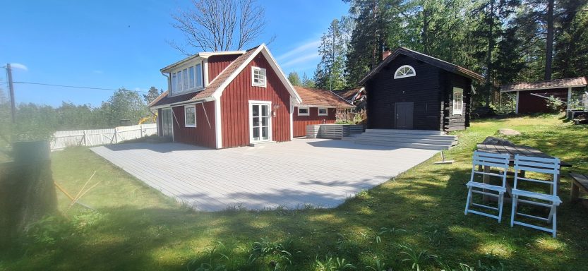 Altan tvått med behandling Röd stuga och garage på en grönskande tomt med däckade ytor och träd.