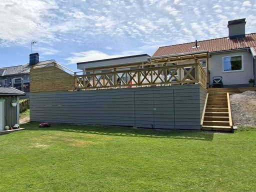Altanbygge med nedsänkt SPA bad En trädgård med en träterrass och angränsande hus under blå himmel.