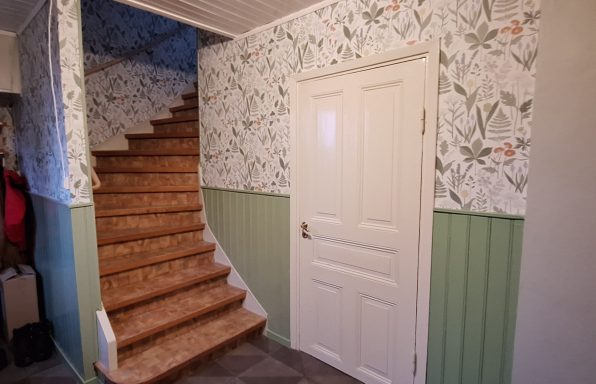 Tapetsering En trappa av trä leder uppåt, omgiven av blommönstrad tapet och en vit dörr.